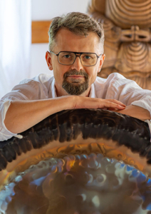 Ein graumelierter Mann mit Brille und Bart und weißem Hemd lehnt mit seinen Unterarmen auf einem Gong
