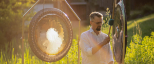 Ein Mann mit grau-meliertem Haar, Bart, Brille und weißem Hemd steht im Grünen bei Sonnenlicht zwischen zwei Gongs. In der rechten Hand hat er einen Schlegel, mit dem er auf einen der Gongs schlägt.