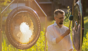 Ein Mann mit grau-meliertem Haar, Bart, Brille und weißem Hemd steht im Grünen bei Sonnenlicht zwischen zwei Gongs. In der rechten Hand hat er einen Schlegel, mit dem er auf einen der Gongs schlägt.