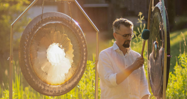Ein Mann mit grau-meliertem Haar, Bart, Brille und weißem Hemd steht im Grünen bei Sonnenlicht zwischen zwei Gongs. In der rechten Hand hat er einen Schlegel, mit dem er auf einen der Gongs schlägt.